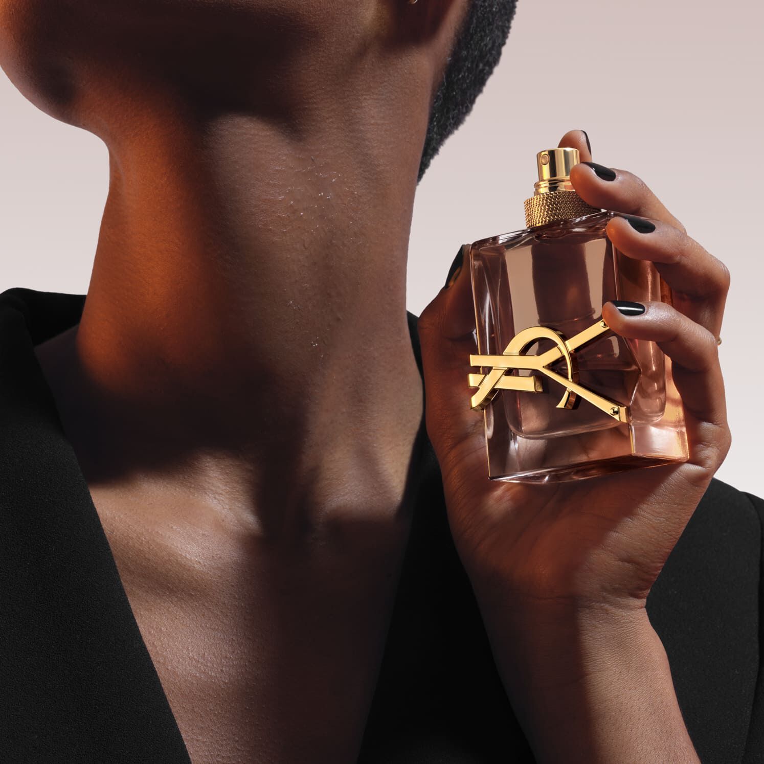Close-up: a hand holds a rectangular glass Libre perfume bottle with a golden YSL logo and cap. The person wears a black blazer. Background is warm orange and white light.​