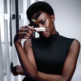 A woman with dark skin and short hair holds Lash Clash Mascara and Loveshine lipstick, partially covering her face, looking at the camera. She wears a black top. Background has reflective surfaces.​