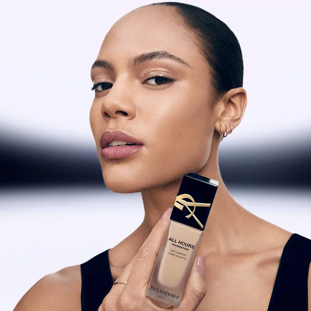 A model with a sleek hairstyle poses confidently, holding a bottle of YSL All Hours foundation in her hand, showcasing a smooth complexion and a subtle makeup look.