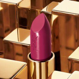 A vibrant berry-colored lipstick with a sleek design sits atop shiny golden geometric shapes. The lipstick tube features the YSL logo.