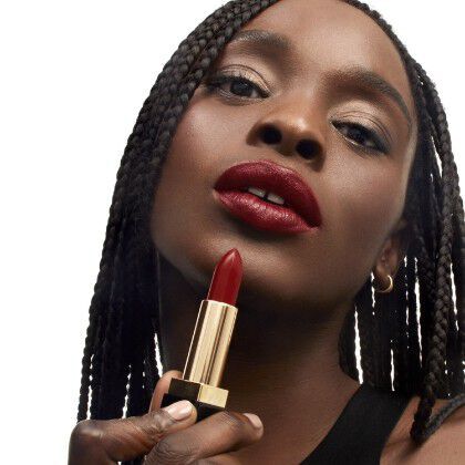 A woman with braided hair holds a red lipstick near her lips, showcasing a bold makeup look against a white background.