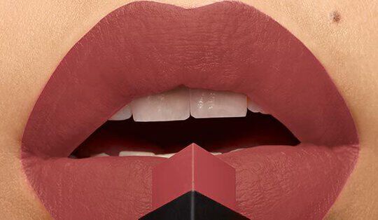 Close-up of a pair of lips painted in a matte, muted reddish-brown shade, with a lipstick bullet positioned at the center.
