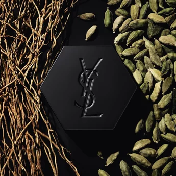 A black hexagonal box with the embossed initials "YSL" is centered on a dark surface, surrounded by dried roots on one side and green cardamom pods on the other.