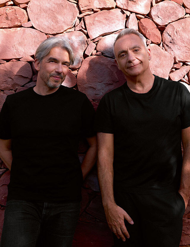 Two men with gray hair, both wearing black t-shirts, stand leaning against a reddish-brown stone wall. The man on the left has a beard and is looking towards the viewer with a slight smile. The man on the right has shorter hair and is also looking towards the viewer, with one hand in his pocket.