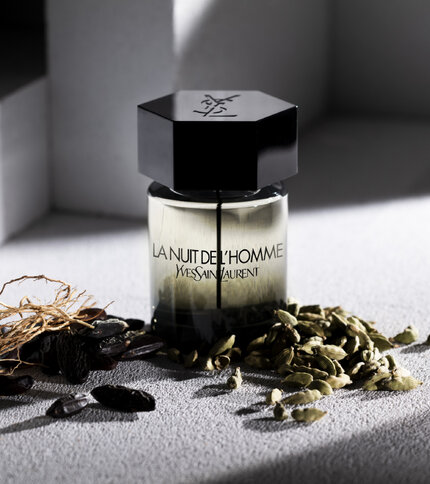 A stylish bottle of "La Nuit de L'Homme" perfume by Yves Saint Laurent, featuring a hexagonal black cap and clear glass body, placed on a textured surface alongside some aromatic spices, including cardamom and other botanicals.
