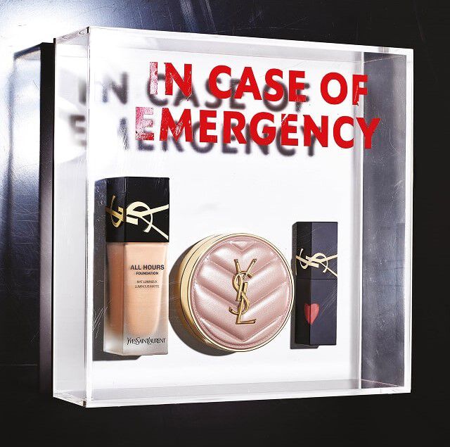 A transparent plexiglass box contains three YSL Beauty makeup products, like an emergency kit. Inside, from left to right, we see a bottle of "All Hours Foundation" liquid foundation, a compact powder or tinted cushion in a pale pink quilted case with the golden YSL logo, and a black lipstick decorated with a small red heart on the cap, also featuring the golden YSL logo. At the back of the transparent box, the text "IN CASE OF EMERGENCY" is written in large red letters, creating a mirror and depth effect, suggesting these products are essentials to always have on hand.