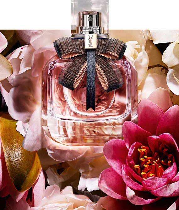 A stylish perfume bottle with a decorative ribbon sits among a backdrop of vibrant flowers, including pink blossoms and soft petals, creating an elegant and floral aesthetic.
