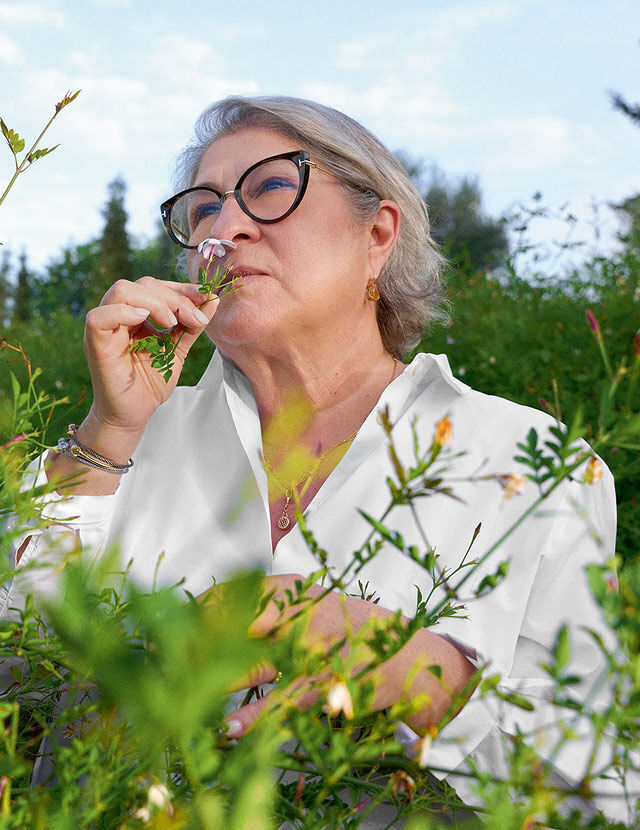 A woman with gray hair and black cat-eye glasses is shown in a lush green garden, smelling a small pink flower held close to her nose. She is wearing a white collared shirt and has jewelry including earrings, a necklace, and bracelets. The sky is bright blue with some clouds visible in the background, suggesting a clear day. The foreground and midground are filled with various green plants and some small, light-colored blossoms.