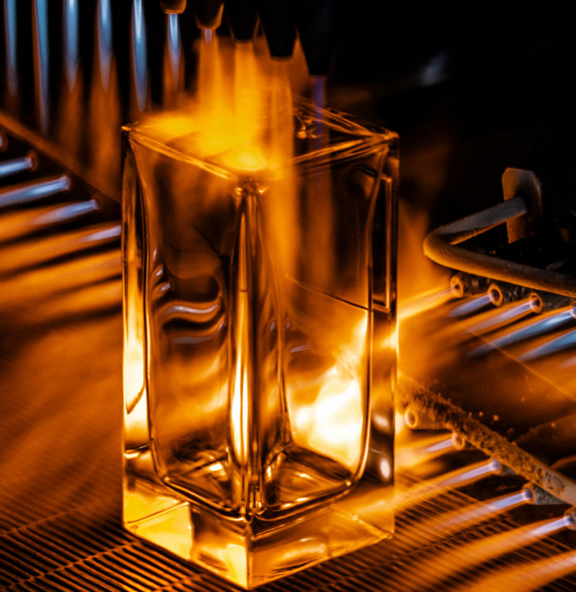A close-up of a clear rectangular perfume bottle being intensively heated by flames from several nozzles, highlighting the industrial process with an orange glow.