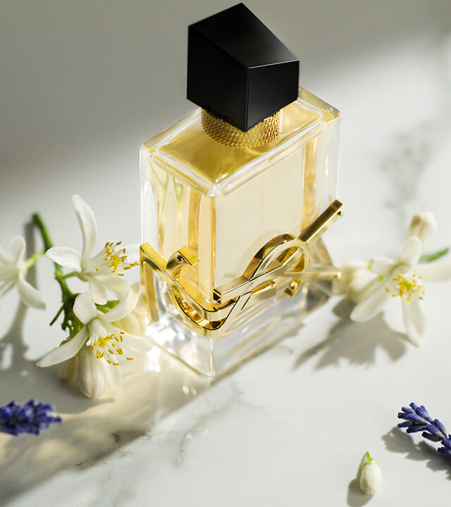 A gold and black perfume bottle is elegantly placed on a marble surface, surrounded by white flowers and lavender sprigs.