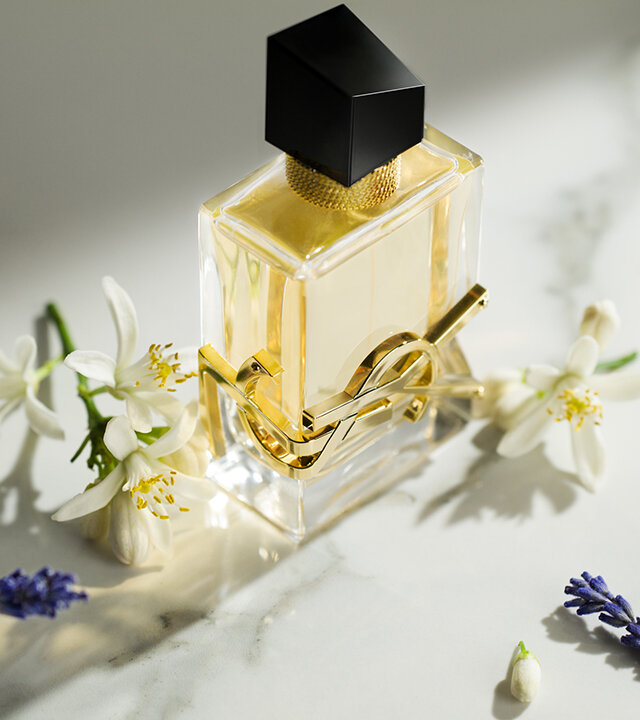 A gold and black perfume bottle is elegantly placed on a marble surface, surrounded by white flowers and lavender sprigs.