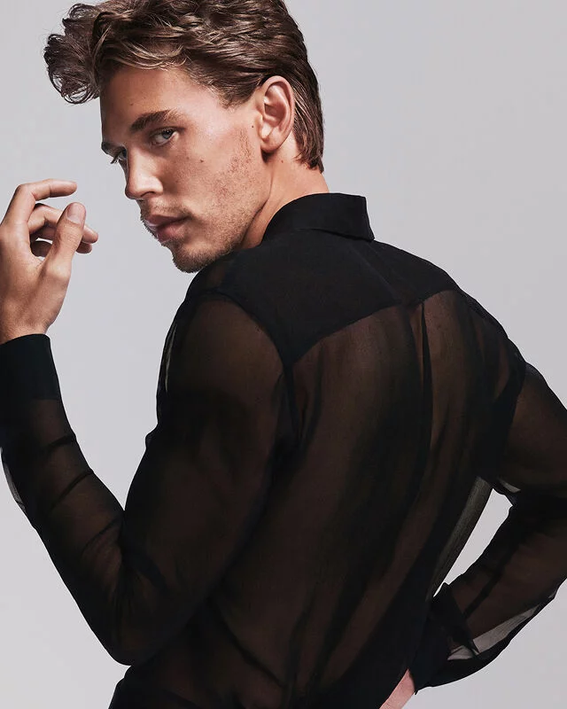 A man with light brown hair and blue eyes, wearing a sheer black shirt, looking over his shoulder.