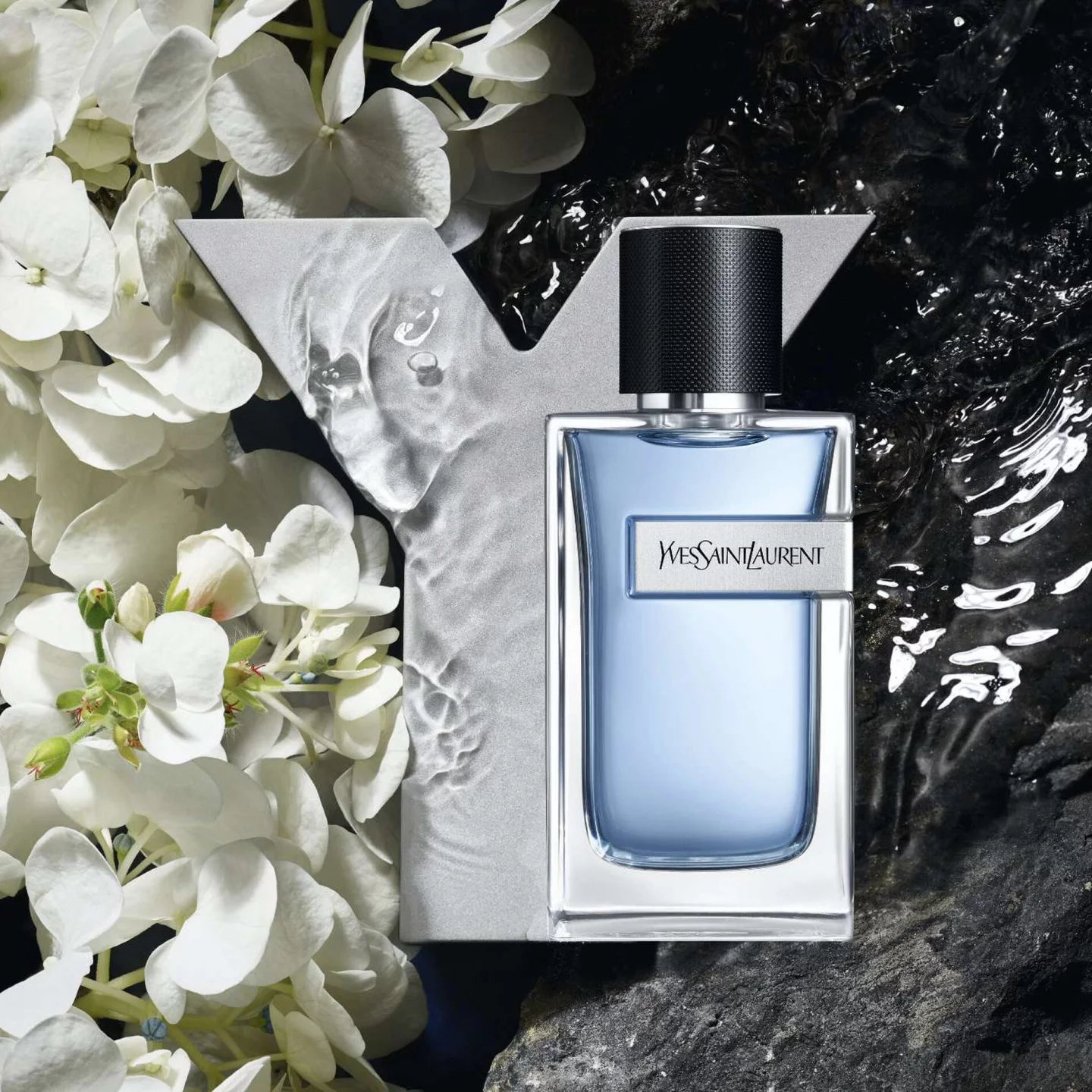 A bottle of Yves Saint Laurent fragrance surrounded by white flowers and water, with a stylized "Y" in the background.