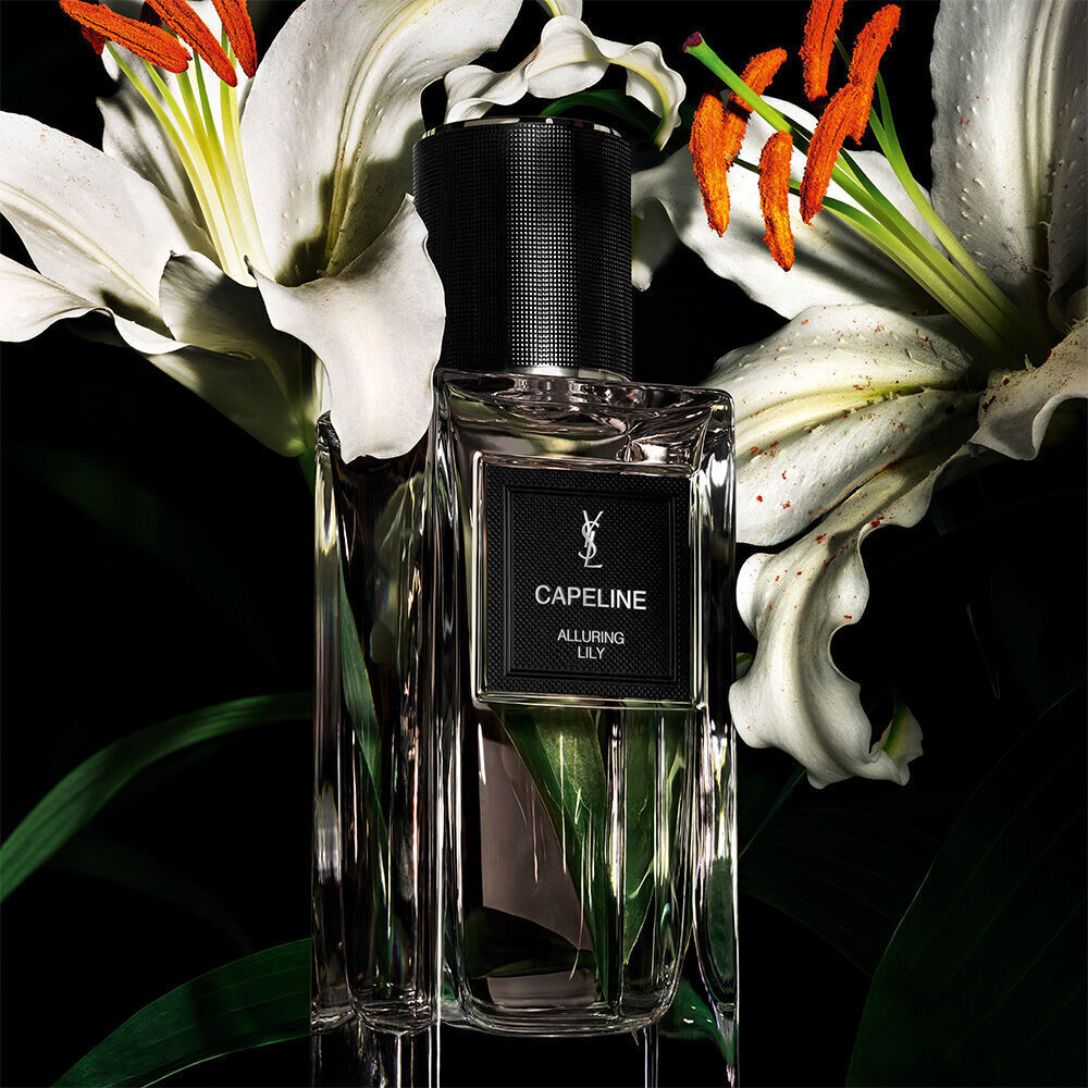 A clear perfume bottle labeled "CAPELINE ALLURING LILY" by YSL, set against a dark background with vibrant white lilies.