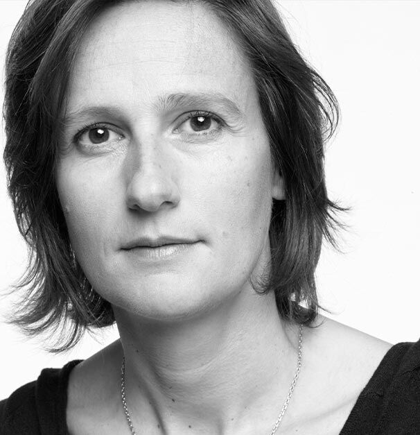 A close-up black and white portrait of a woman with shoulder-length hair, looking directly at the camera with a neutral expression.