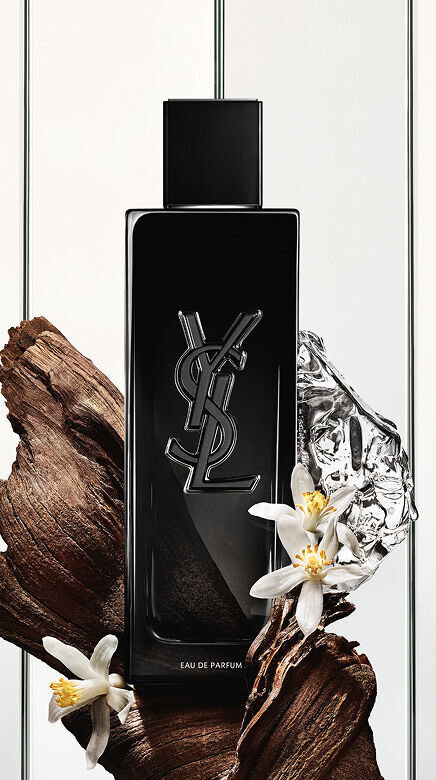 A black Yves Saint Laurent Eau de Parfum bottle with the YSL logo, surrounded by dark wood pieces, white delicate flowers, and a crumpled silver reflective object.