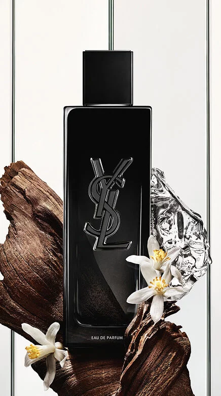 A black Yves Saint Laurent Eau de Parfum bottle with the YSL logo, surrounded by dark wood pieces, white delicate flowers, and a crumpled silver reflective object.