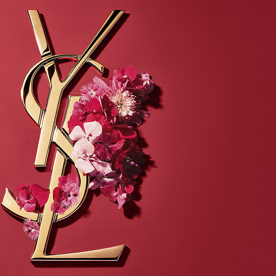 A large, shimmering pink glitter heart on a soft pink background. The iconic gold Yves Saint Laurent (YSL) monogram logo is elegantly overlaid vertically across the heart.​