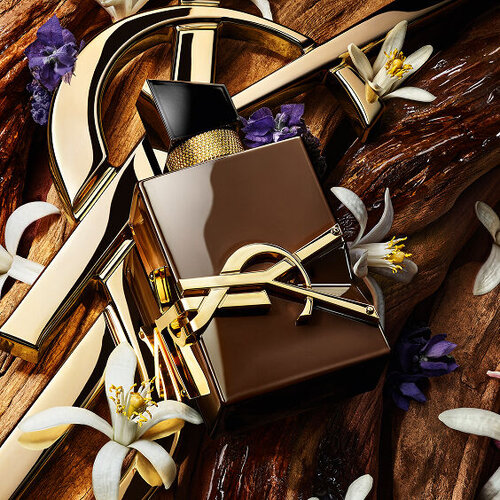 YSL Libre perfume bottle surrounded by gold hardware, white orange blossoms, and purple flowers on a wooden background.