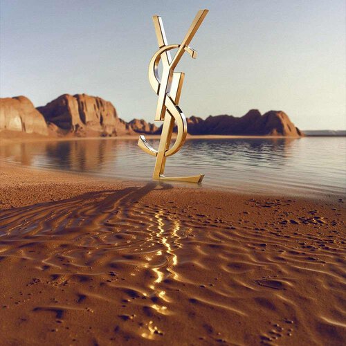 A large, iconic 3D gold YSL (Yves Saint Laurent) logo standing on a desert shoreline. The polished gold monogram reflects the sunlight and is positioned in front of a calm body of water with dramatic red rock formations and desert mountains in the background under a clear sky.