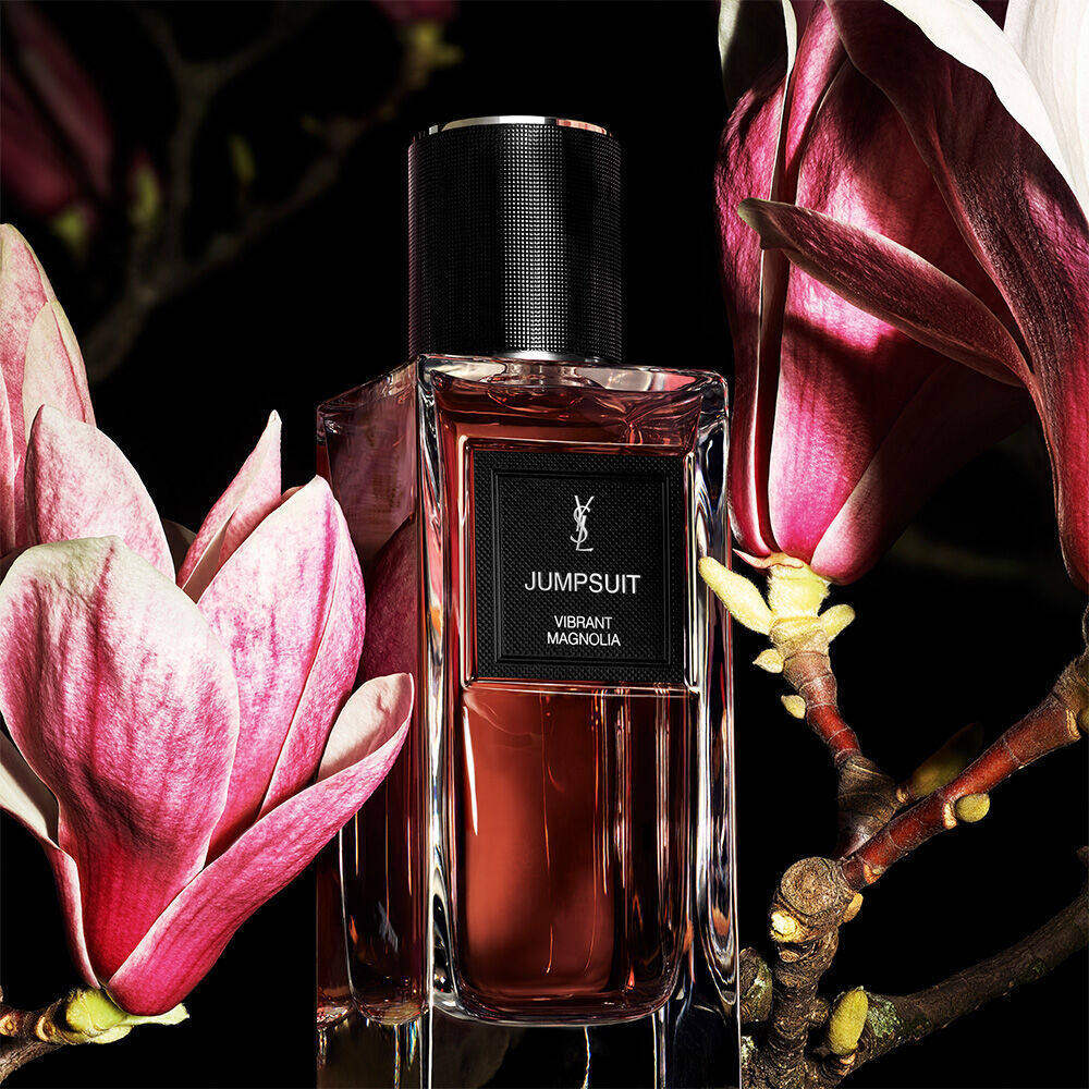 A reddish-brown perfume bottle labeled "JUMPSUIT VIBRANT MAGNOLIA" by YSL, set against a dark background with large, pink magnolia petals.