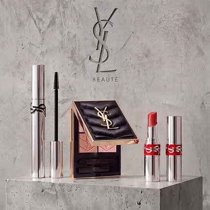 Promotional display of YSL Beaute makeup products including mascara, a quilted eyeshadow palette, and lipsticks against a concrete background.