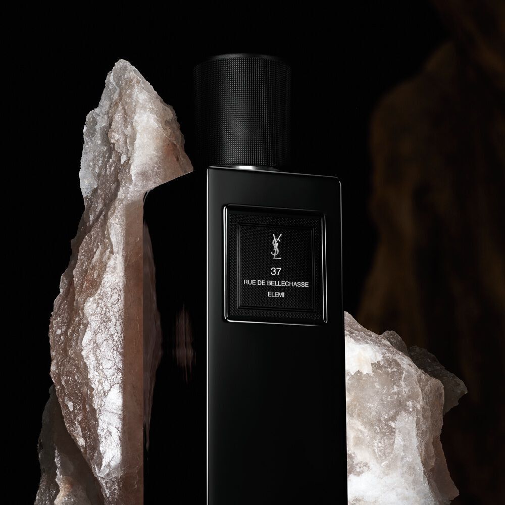A sleek, rectangular black perfume bottle labeled "37 RUE DE BELLECHASSE ELEMI" by YSL, framed by large, rough, white crystal formations.
