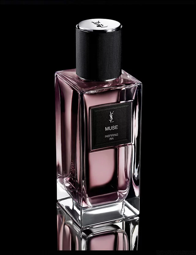 A rectangular, pink-tinted perfume bottle labeled "MUSE INSPIRING INK" by YSL, with a black textured cap, sits on a reflective surface against a dark background.