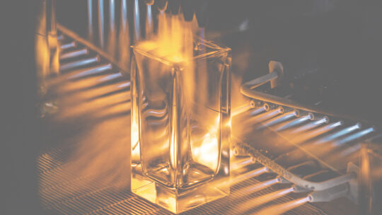 A clear rectangular perfume bottle is shown being heated by flames, likely part of a glass-making or treatment process.