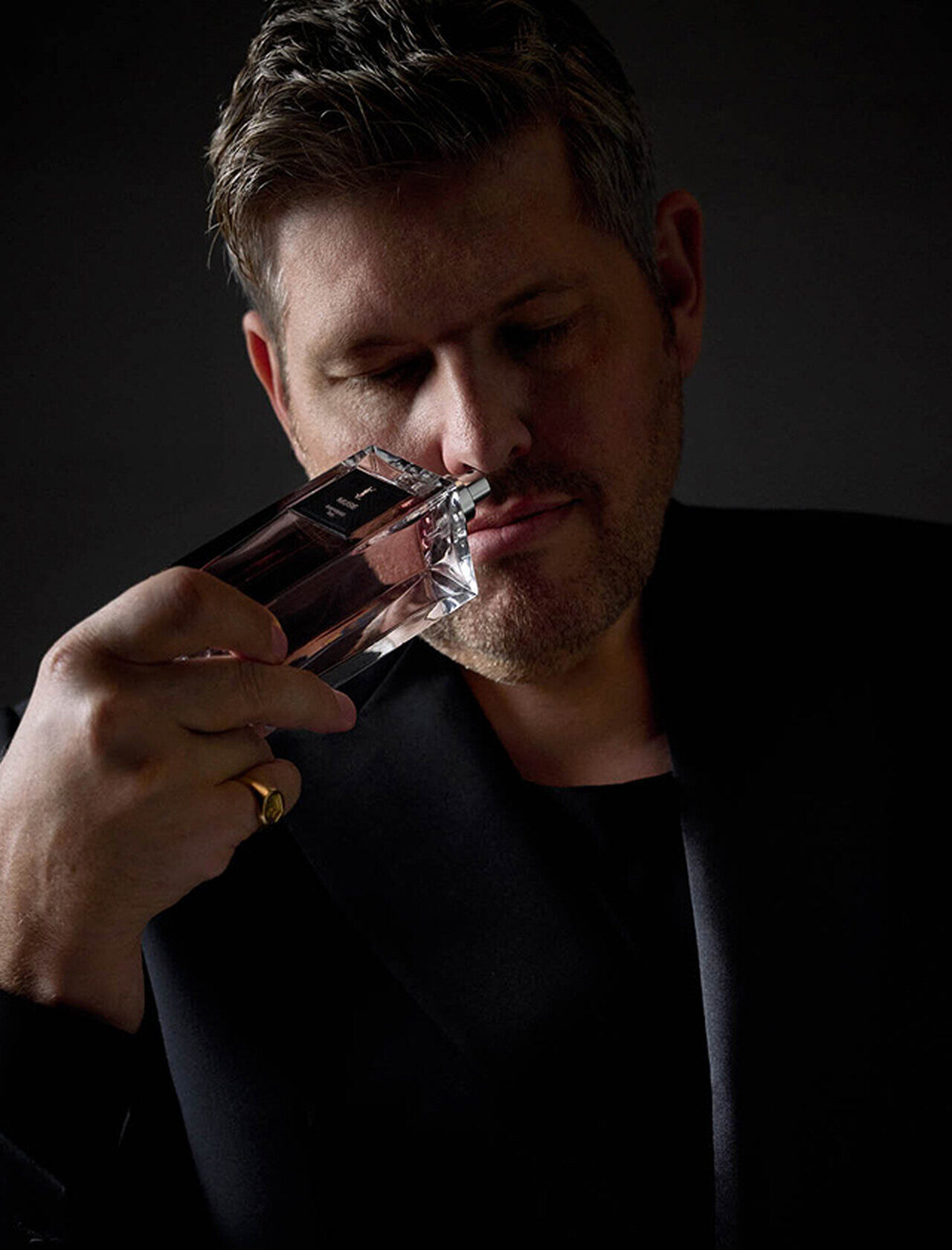 A man in a dark top holds a perfume bottle to his nose, deeply inhaling its scent.