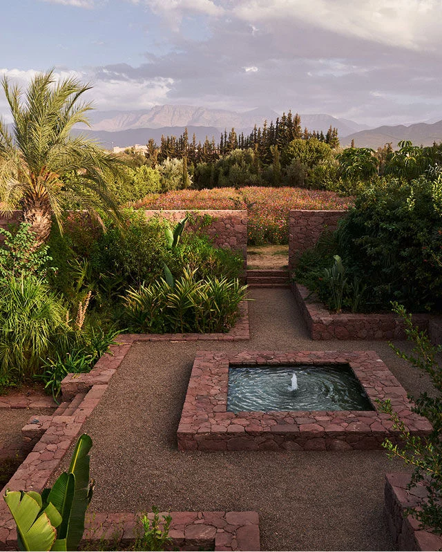 A verdant garden features a square stone fountain with bubbling water in the foreground, surrounded by gravel paths and low stone walls. Lush green plants and shrubs fill the garden beds, with a field of red and pink flowers in the middle ground. In the background, mountains rise under a soft, cloudy sky, and a large palm tree stands prominently on the left.