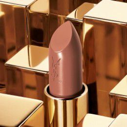 A close-up image of a nude lipstick with a polished golden casing, set against a backdrop of shiny gold geometric shapes. The lipstick features the YSL logo.