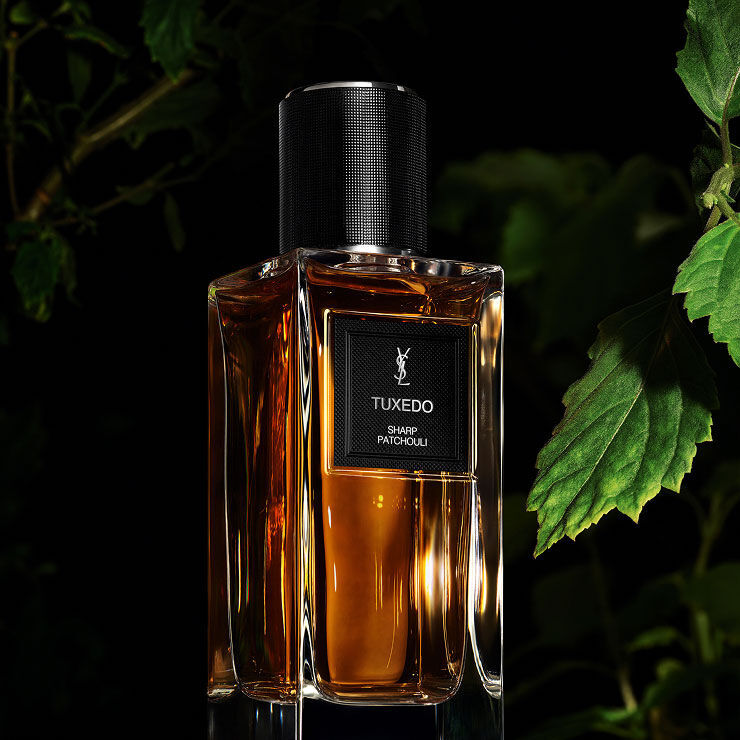 A golden-toned perfume bottle labeled "TUXEDO SHARP PATCHOULI" by YSL, with soft green foliage providing a natural background.