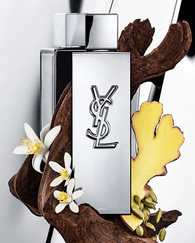 YSL Y L'ABSOLU perfume bottle surrounded by dark wood, white orange blossom flowers, ginger, and cardamom pods.