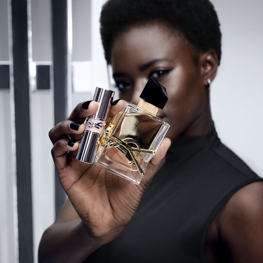 A woman with dark skin and short hair holds up a bottle of Yves Saint Laurent perfume and a coordinating lipstick, both featuring the iconic YSL logo.