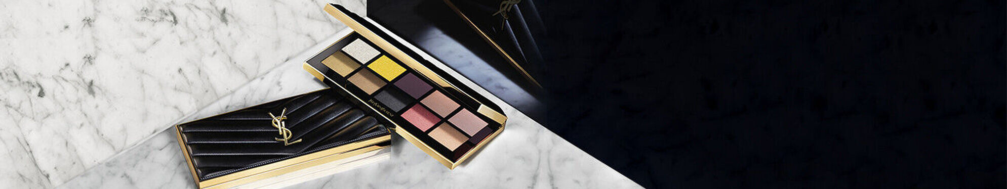 An elegant black eyeshadow palette with a gold YSL logo lies open on a marble surface, revealing a range of eye shadow shades including neutrals, purples, and yellows.