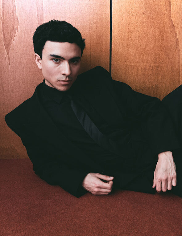 A young man with dark, styled hair poses in a black suit against a wooden background, giving a serious expression.