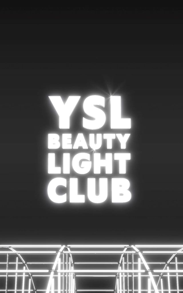 A wide, dark shot reveals the "YSL BEAUTY LIGHT CLUB" logo in luminous white letters, floating in the center of a black space. Below the logo, a complex structure of metallic bars or glowing white neon lights creates a grid or stage effect, suggesting a luminous installation or a futuristic club environment. The overall impression is one of a modern and chic atmosphere.