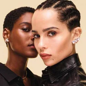 Two women with distinct hairstyles and styles are posing closely together against a soft, pastel background. One has short, dark hair and a confident expression, while the other has long, slicked-back hair and is wearing a leather jacket.