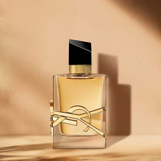 A clear glass perfume bottle with golden liquid, a black and gold cap, and a prominent gold YSL logo, set against a warm, neutral background with soft shadows.