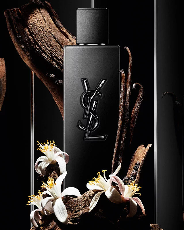 Black YSL perfume bottle surrounded by wood, vanilla pods, and white flowers.