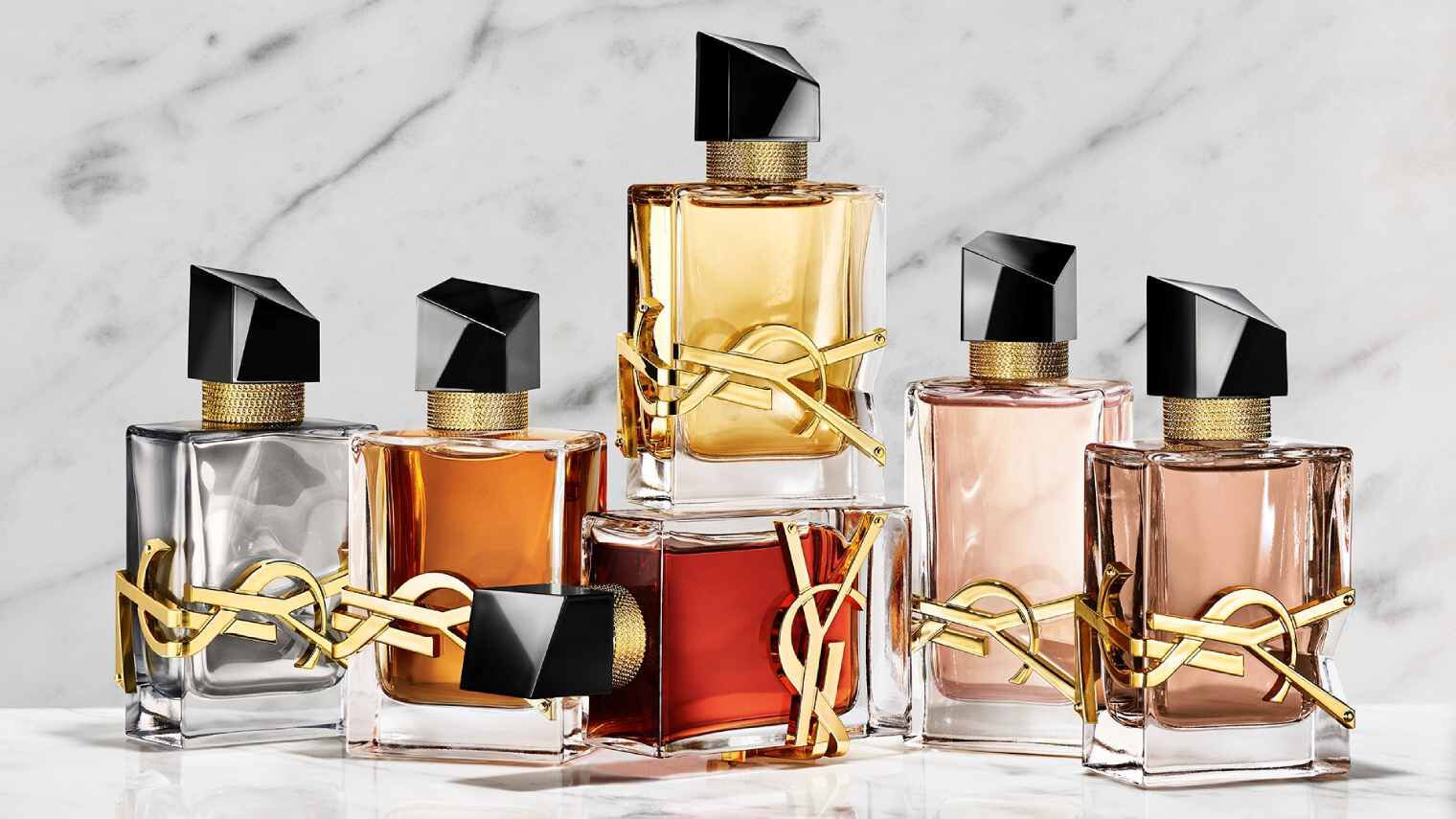 A hand holds a red YSL perfume bottle next to two other fragrances on a stone base.