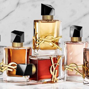 A hand holds a red YSL perfume bottle next to two other fragrances on a stone base.