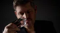 A man in a dark top holds a perfume bottle to his nose, deeply inhaling its scent.