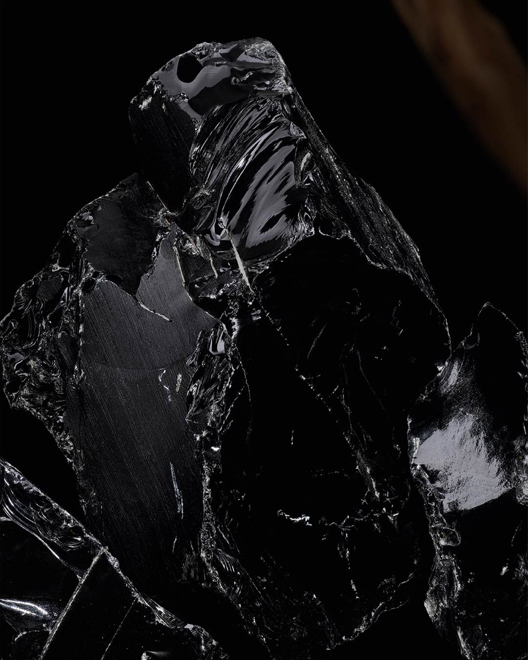 A close-up view of glossy black obsidian rock with a smooth, reflective surface, set against a dark background. A piece of natural wood is partially visible on the side.