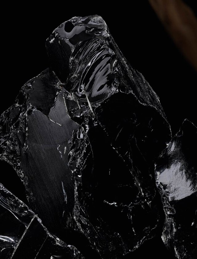 A close-up view of glossy black obsidian rock with a smooth, reflective surface, set against a dark background. A piece of natural wood is partially visible on the side.