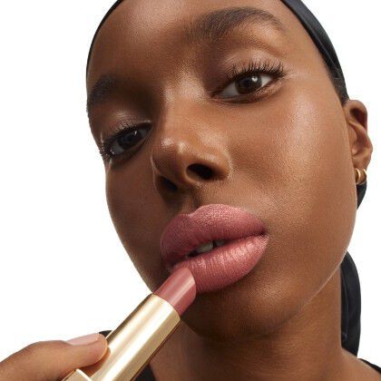 A close-up portrait of a woman applying nude lipstick, showcasing her full lips and glowing skin against a white background. She has a black headscarf and subtle makeup.
