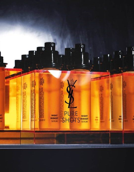 Row of amber-colored Yves Saint Laurent Pure Shots Night Reboot Serum bottles with black pump tops. The YSL logo is prominent on each bottle, which stands on a reflective surface against a dark, smoky background, highlighting the product line.