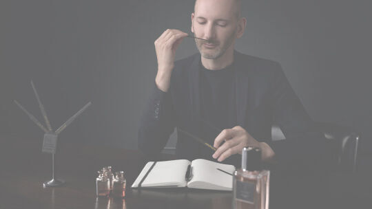 A perfumer in a dark suit attentively smells a perfume blotter in a dimly lit setting.