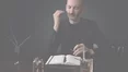 A perfumer in a dark suit attentively smells a perfume blotter in a dimly lit setting.